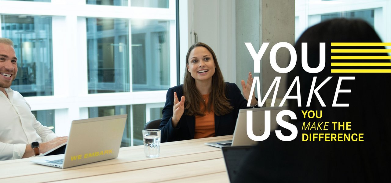 Three colleagues sit across from each other laughing at a meeting table with laptops. Above the picture is the "You Make Us" lettering.